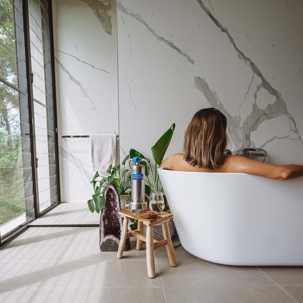A woman sits in a white freestanding bathtub in a modern bathroom with large marble-look walls and floor tiles. A small wooden stool with a water filter bottle stands beside the tub near a window with a view of greenery outside.