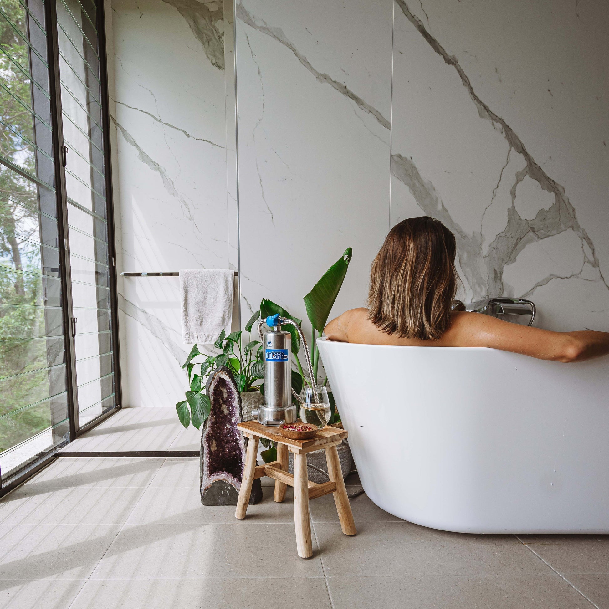 A woman sits in a white freestanding bathtub in a modern bathroom with large marble-look walls and floor tiles. A small wooden stool with a water filter bottle stands beside the tub near a window with a view of greenery outside.