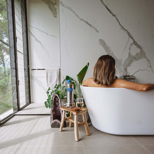 A woman sits in a white freestanding bathtub in a modern bathroom with large marble-look walls and floor tiles. A small wooden stool with a water filter bottle stands beside the tub near a window with a view of greenery outside.