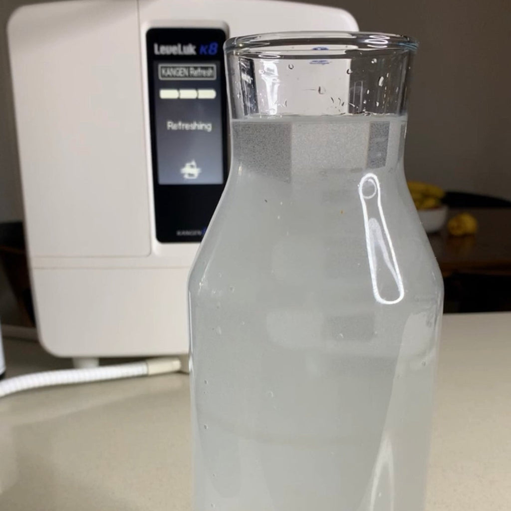 A clear glass water bottle filled with Kangen water, placed on a countertop in front of a white water dispenser.