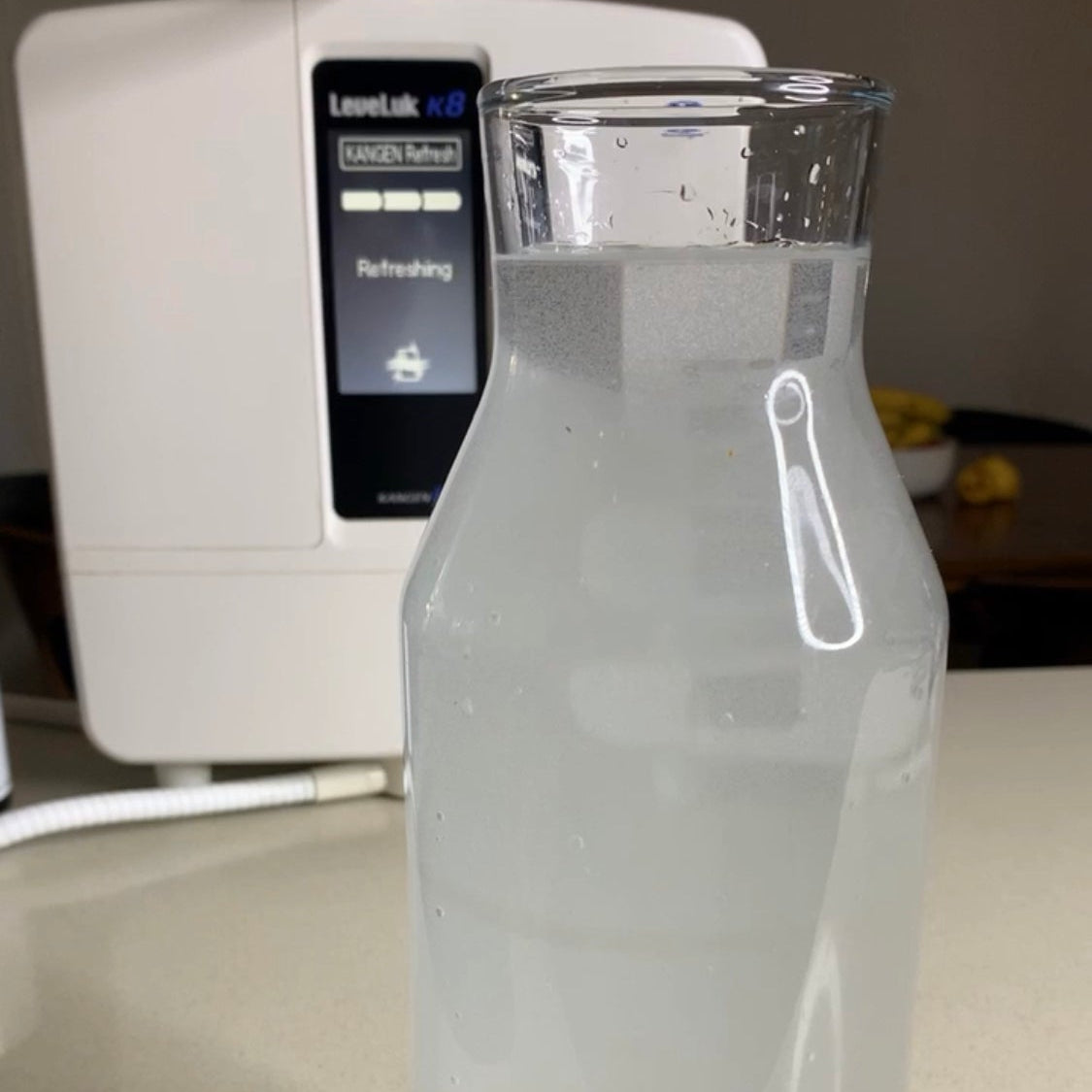 A clear glass water bottle filled with Kangen water, placed on a countertop in front of a white water dispenser.